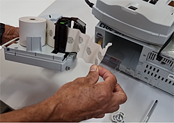 Imagem exibindo o módulo impressor. Uma mão está fazendo o ajuste do papel neste módulo.