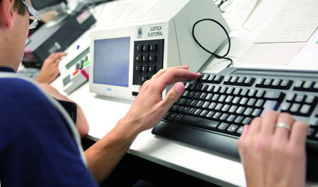 Imagem de um homem digitando em um teclado de computador e ao lado há a imagem de uma urna eletrônica.