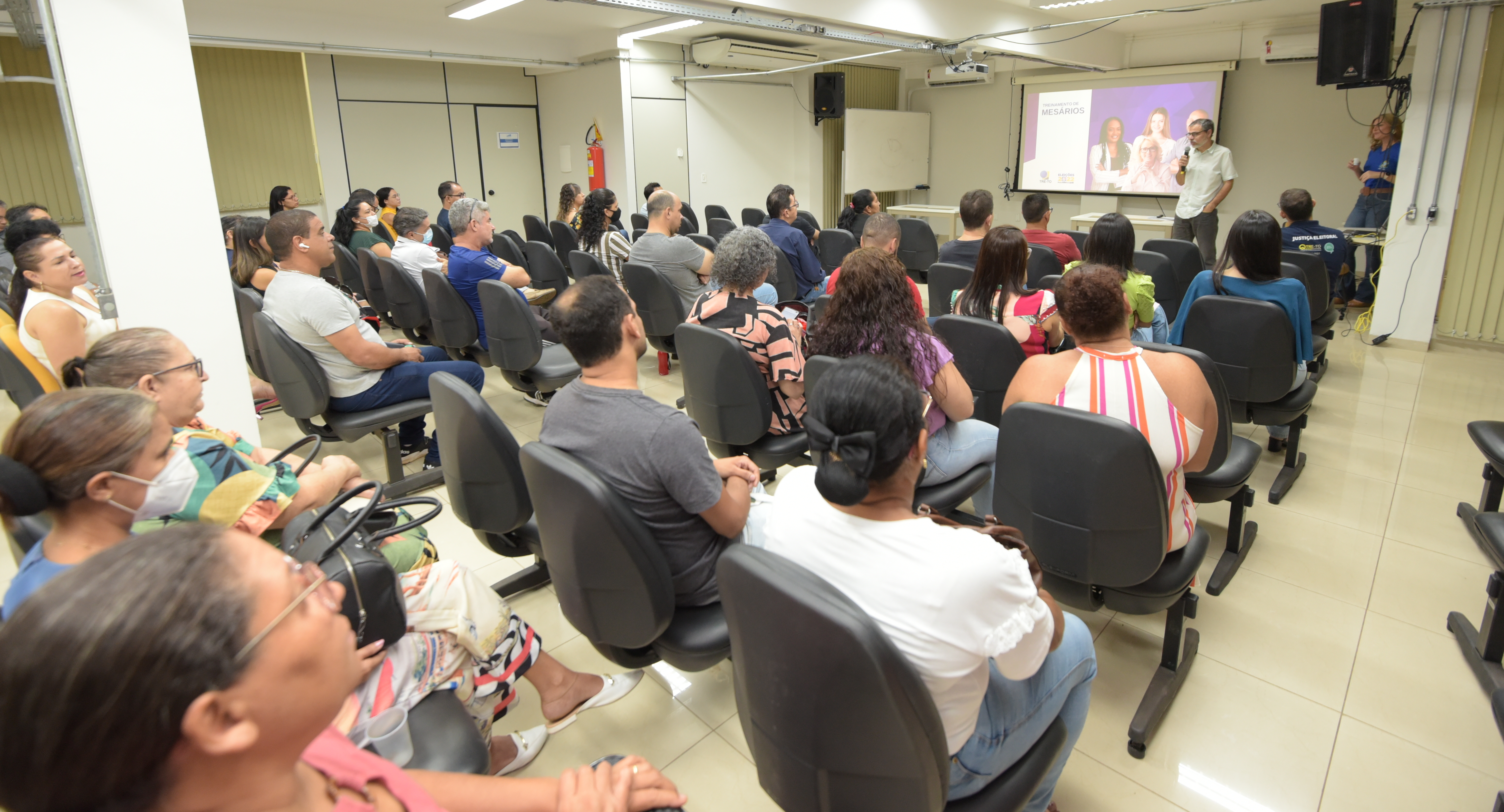 Foto de um treinamento de mesários com um servidor 
dando palestras para um grupo de mesários, num auditório